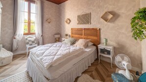 A bedroom with a wooden ceiling, a large bed with white bedding, a bedside table with a lamp, a wall-mounted fan, and a window with curtains. - Charming house with AC, pets OK - 4 guests (Parentis-en-Born)