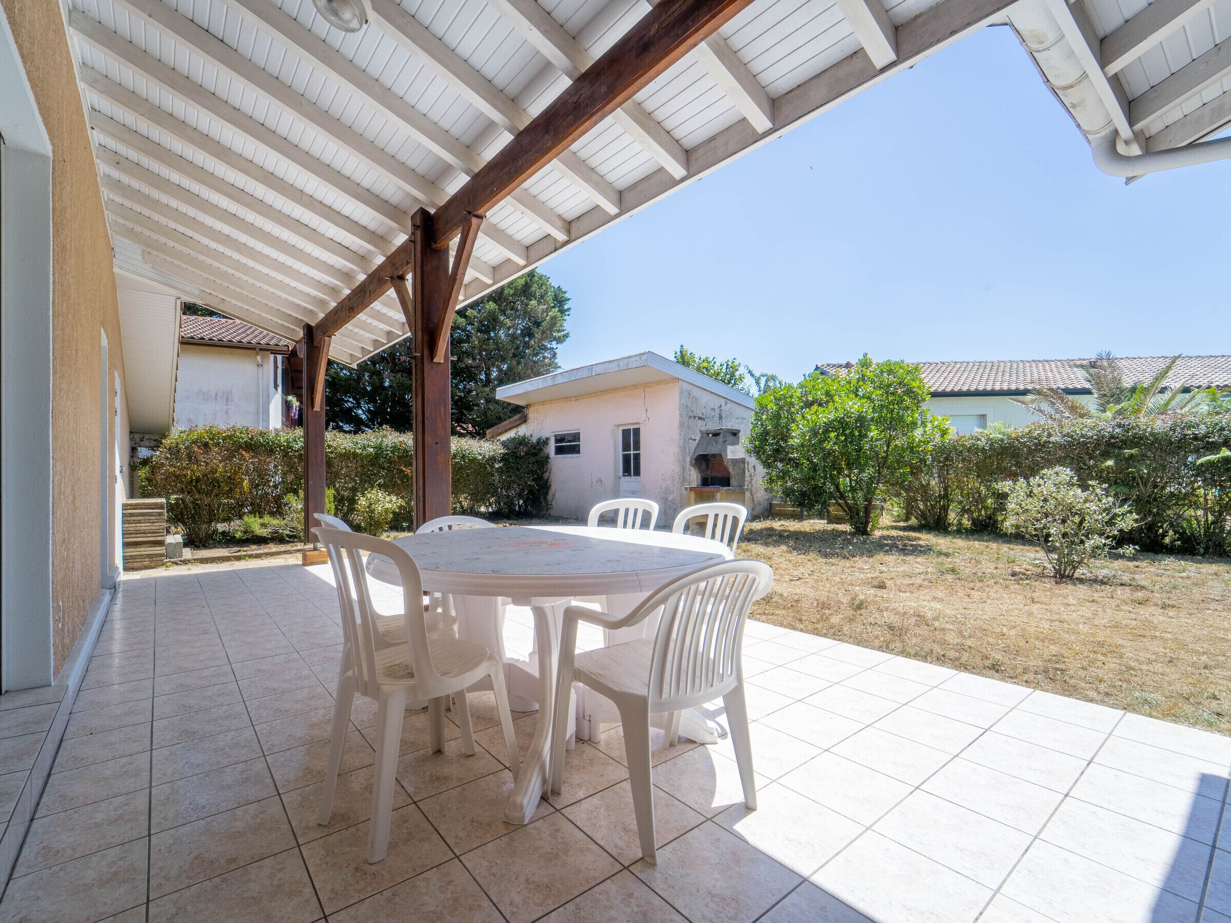 Une terrasse couverte avec une table et des chaises, un sol carrelé et une vue sur une maison et la verdure.