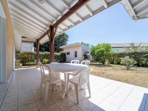 Eine überdachte Terrasse mit Tisch und Stühlen, ein Fliesenboden und Blick auf ein Haus und Grünflächen.