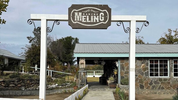 En rustik entré med en skylt som visar texten ”RANCHO MELING”, en gång som leder till en byggnad med en fasad av sten och ett tak av metall.