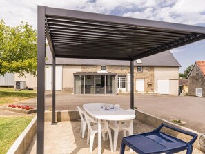 A covered outdoor seating area with a table and chairs, a building with a stone facade in the background, and a clear sky. - Spacious house with parking - 6 guests (Les Loges-Marchis)