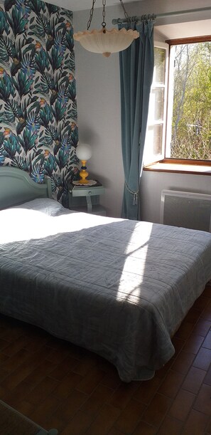 Ein Schlafzimmer mit Bett, Nachttisch mit Lampe, Fenster mit Vorhängen und Heizkörper.