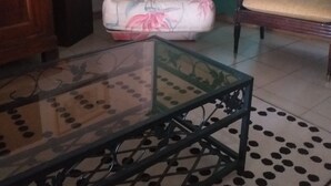 A room with a glass-top table, a floral armchair, a sofa with a patterned cushion, a wooden cabinet, and a green door. - A CHARACTERFUL PROVENÇAL FARMHOUSE FOR 2 TO 10 VACATIONERS. (Goudargues)