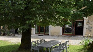 Ein Sitzbereich im Freien mit Tisch und Stühlen unter einem Baum, ein Steinhaus mit einer Tür im Hintergrund.