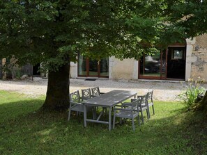 Ein Sitzbereich im Freien mit Tisch und Stühlen unter einem Baum, ein Steinhaus mit einer Tür im Hintergrund.