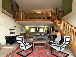 A spacious living area with a high ceiling, a wooden staircase, a kitchen in the background, and a fireplace.