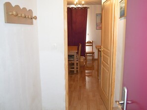 Ein schmaler Flur mit Holzboden, einem Holz-Kleiderständer, einem violetten Vorhang und einer Tür zu einem Zimmer mit Bett und Sessel.