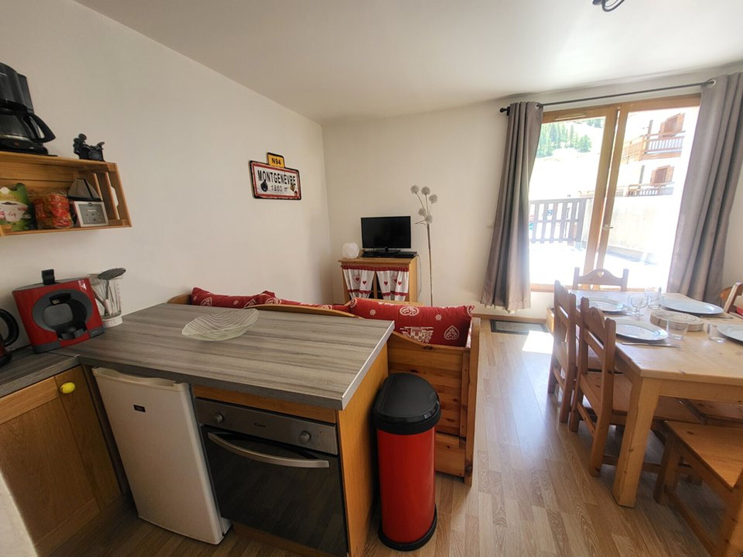 A compact kitchen with a refrigerator, stove, and a coffee machine. There is a dining area with a table set for four, and a TV on a small stand.