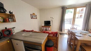 Una cocina compacta con refrigerador, estufa y cafetera. Hay una zona de comedor con una mesa dispuesta para cuatro personas, y un televisor sobre un pequeño mueble.