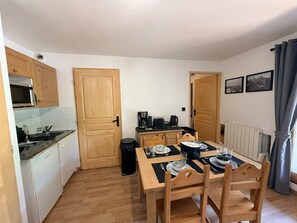 A compact kitchen with wooden cabinets, a microwave, and a dining area with a table set for two. - Cozy apartment at the foot of the slopes - 2 guests (Montgenèvre)