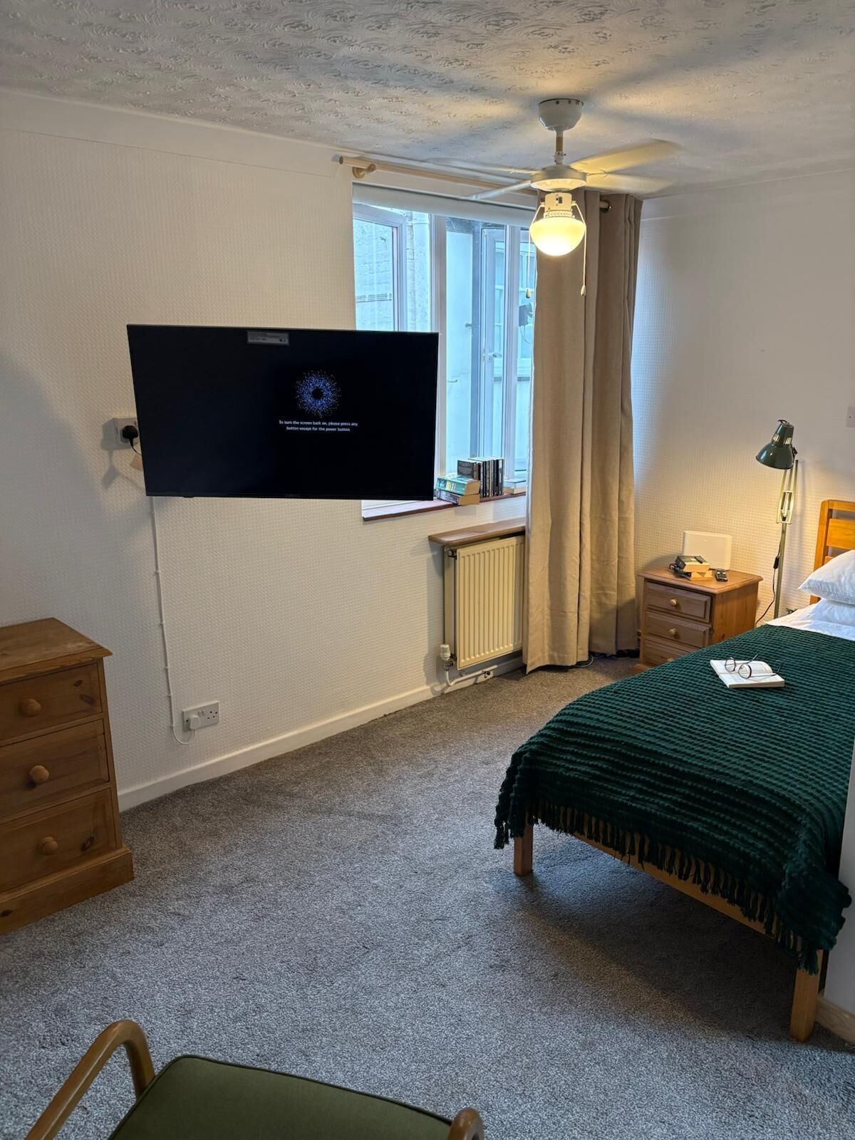A bedroom with a bed, a desk, a chair, a TV mounted on the wall, a window with curtains, and a radiator.