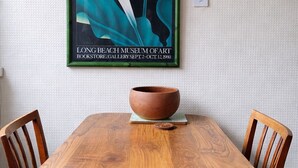 Uma mesa de jantar com uma tigela e cadeiras, uma obra de arte emoldurada acima e um interruptor de luz na parede.