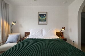 Ein Hotelzimmer mit einem Bett, zwei Nachttischlampen, einem Sessel, einem gerahmten Kunstwerk an der Wand und einer grün gemusterten Decke.
