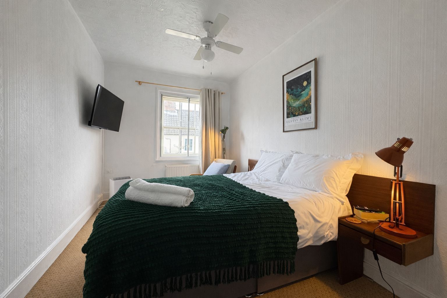 Een slaapkamer met een bed, een plafondventilator, een wandtv, een bedlampje, een raam met gordijnen en een lijst met een afbeelding aan de muur.