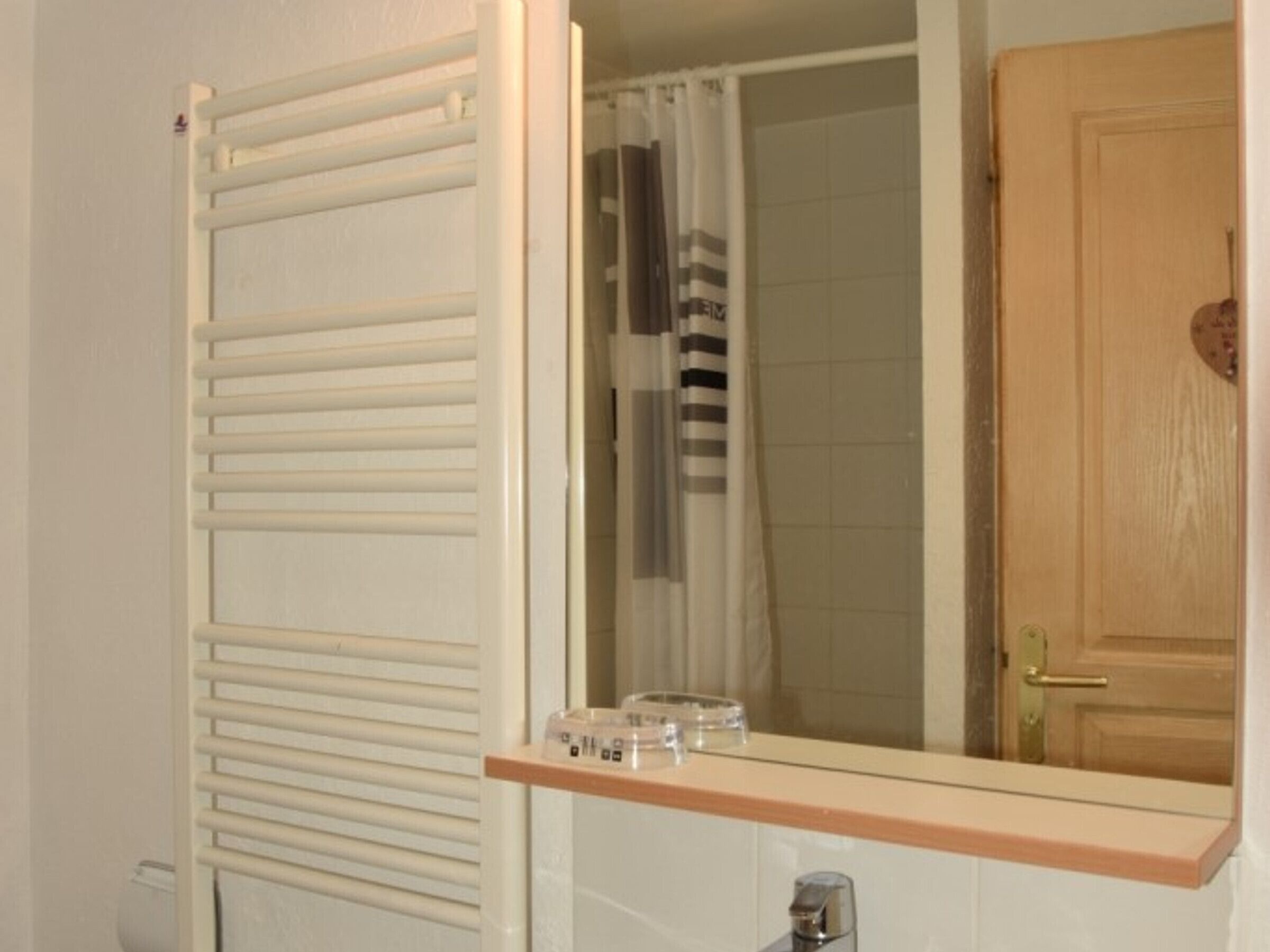 A bathroom with a white radiator, a shower curtain, a wooden door, and a mirror with a shelf above it.