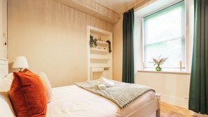 Una cama bien hecha con almohadas, una lámpara de noche, un estante con adornos y una ventana con cortinas.