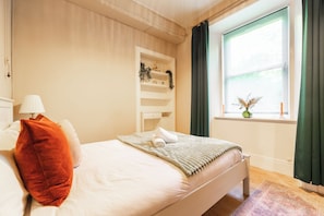 A neatly made bed with pillows, a bedside lamp, a shelf with decorative items, and a window with curtains. - Modern 1BR Walk to Haymarket & Edinburgh Castle (Edinburgh)