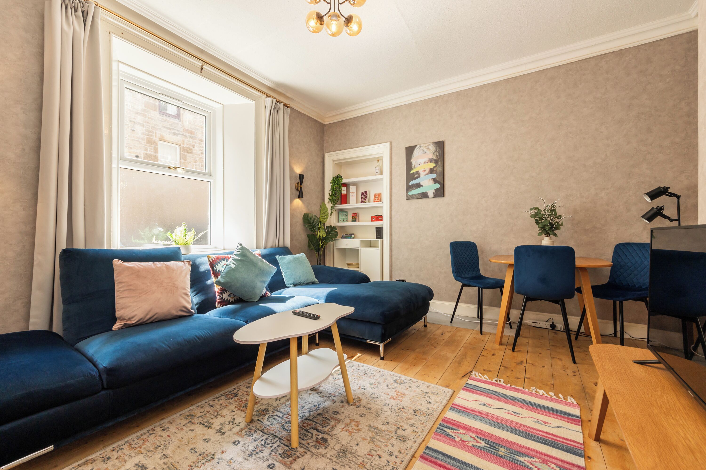 Un salon moderne avec un canapé bleu, une petite table blanche, une table basse en bois, un tapis à motifs, une bibliothèque et un coin repas avec des chaises.