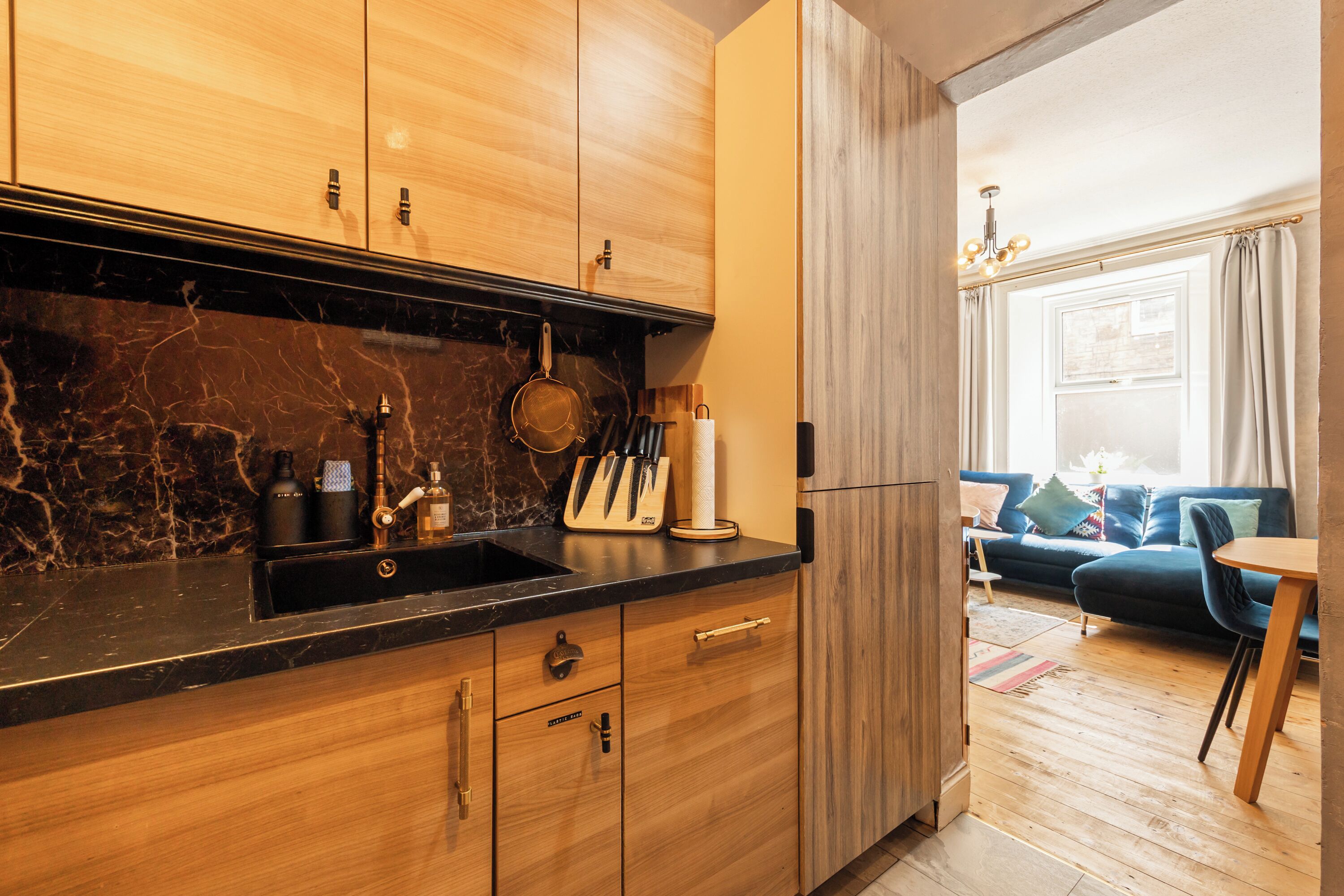 A modern kitchen with wooden cabinets, a marble countertop, and a view into a living area with a sofa and a table.