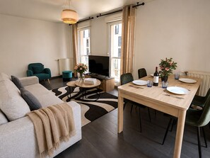 Un salon moderne avec un canapé, une table à manger dressée pour deux personnes, un téléviseur et une fenêtre avec des rideaux.