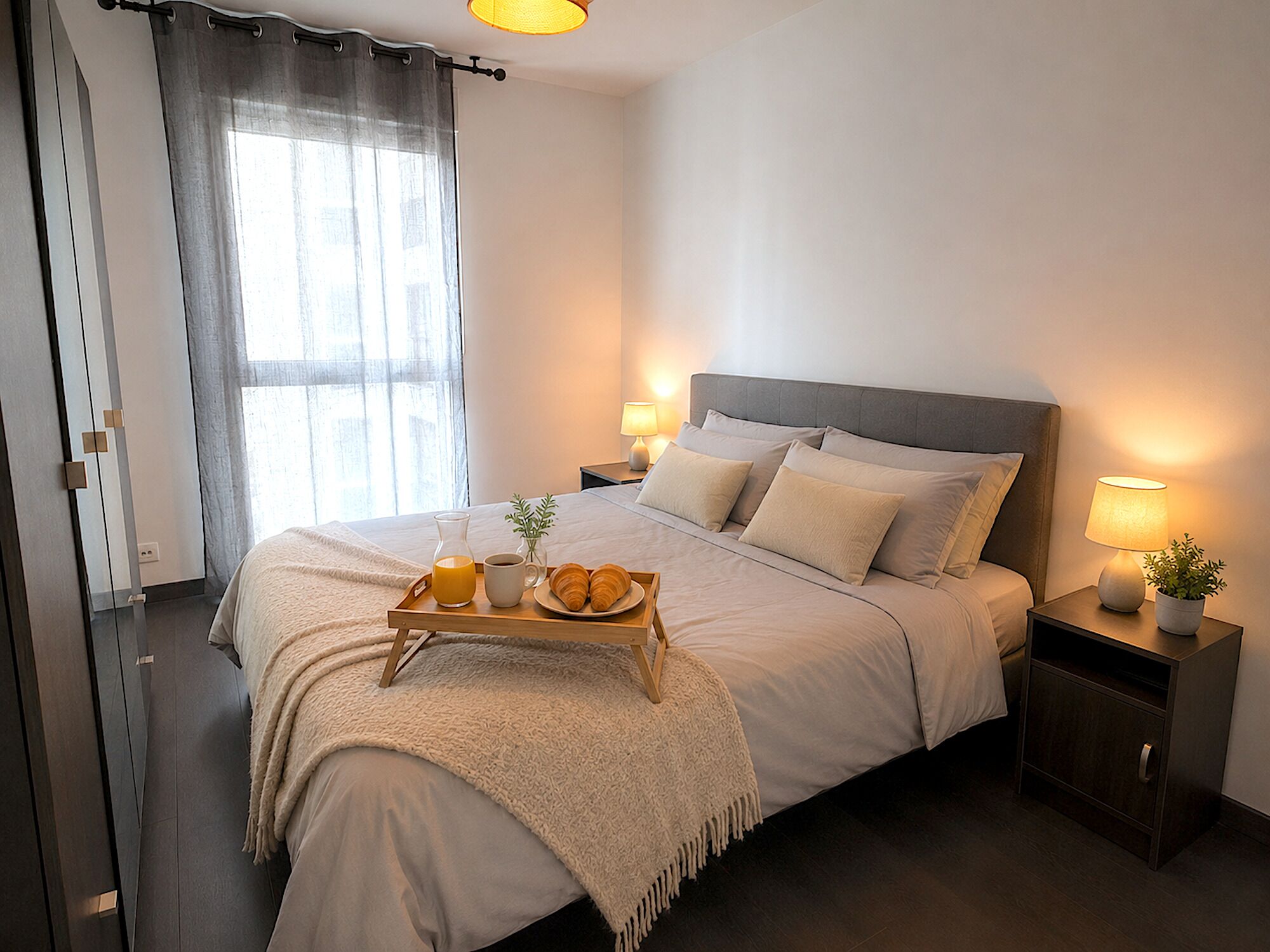 Una cama bien hecha con almohadas, una mesita de noche con lámpara, una bandeja con cruasanes y una bebida, y una ventana con cortinas.