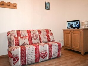 Ein Sofa mit einer rot-weißen Musterdecke, ein Holzschrank mit einem Fernseher und ein gerahmtes Bild an der Wand.