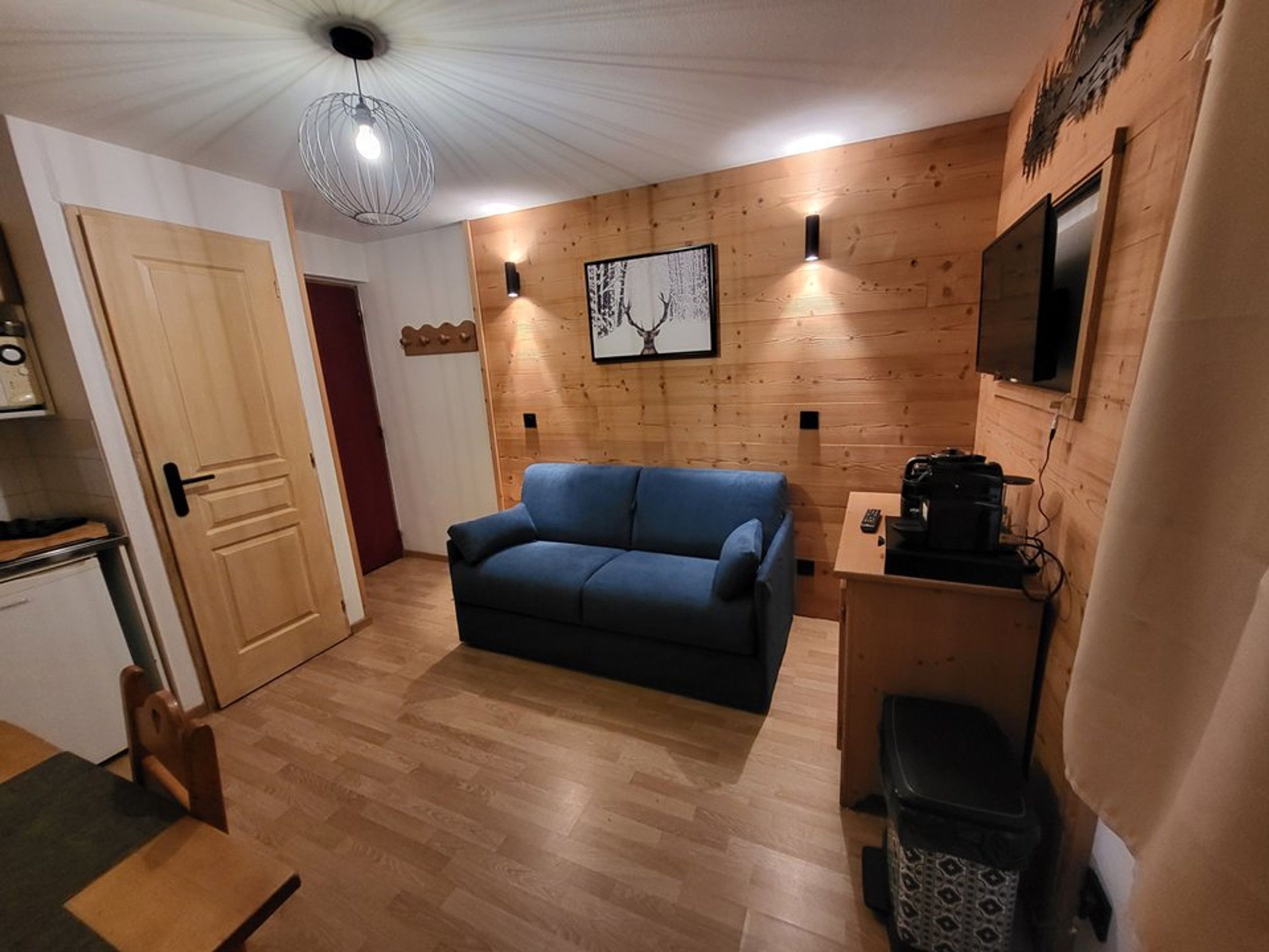 Une pièce avec un canapé bleu, des murs en bois, un luminaire au plafond, un téléviseur fixé au mur et une petite table sur laquelle se trouve une machine à café.