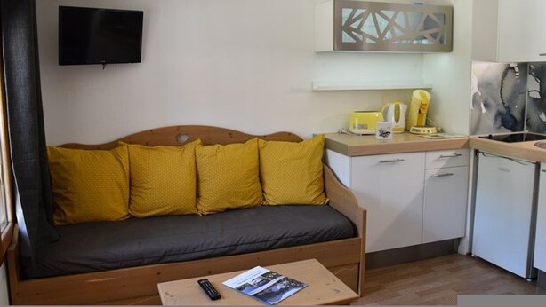 A compact room with a sofa, a small table with a remote control and magazine, a kitchen area with a sink and a microwave, and a flat-screen TV mounted on the wall. - Studio cabin facing slopes - Pets OK (Montgenèvre)
