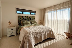 Ein Schlafzimmer mit einem Bett, Nachttischen, einem Fenster mit Jalousien und Blick ins Freie.