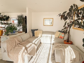 A living room with a sectional sofa, multiple potted plants, a framed picture on the wall, and a window allowing natural light.