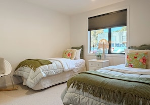 A bedroom with two single beds, a window with blinds, a bedside table with a lamp, and a chair.