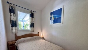 Ein Schlafzimmer mit einem Bett, einem Nachttisch, einem Fenster mit Vorhängen, einem an der Wand hängenden Bild und einem Holzboden.