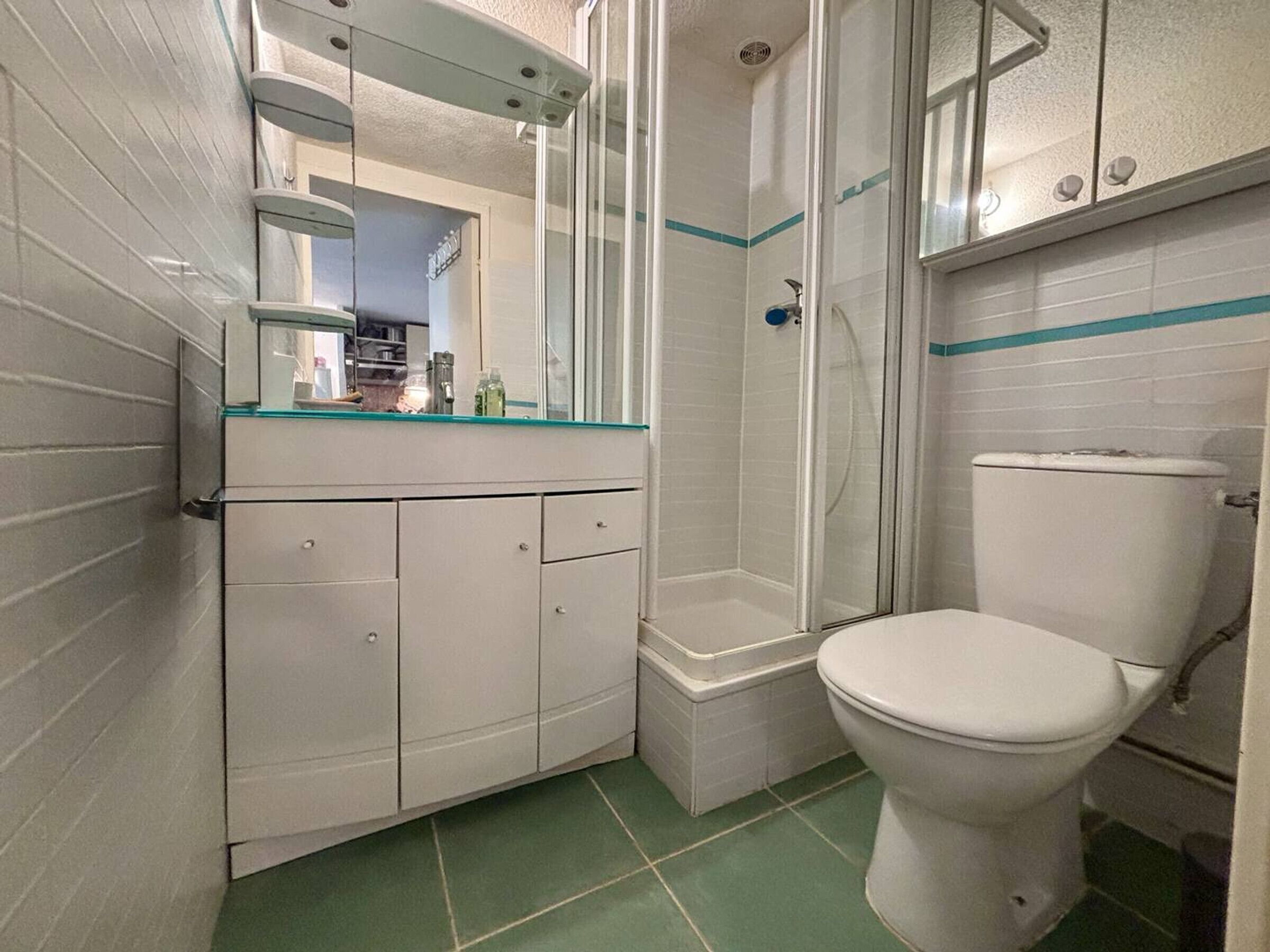 Casa de banho com sanita branca, cabine de duche em vidro e parede revestida a mosaico branco com uma faixa verde.