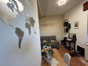 A living room with a world map mural, a grey sofa, a wooden dining table with a lace runner, and a bookshelf with plants. - Teresa's Holiday Home | Your Gateway to Western Sicily with private parking (Salemi)