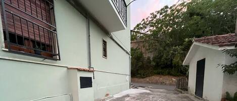 A narrow alleyway with a concrete path, a small building with a black door, and a window with metal bars.