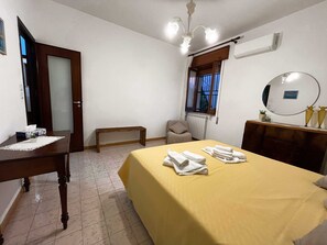 A hotel room with a bed, a bedside table, a chair, a desk, and a window with curtains. - Teresa's Holiday Home | Your Gateway to Western Sicily with private parking (Salemi)