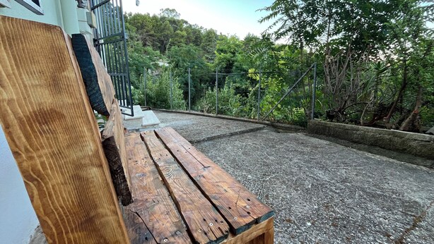 Una panca di legno di fronte a un edificio, con un vialetto sterrato che conduce a un'area giardino recintata.
