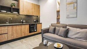 A modern kitchen with wooden cabinets and a built-in oven, a grey sofa with cushions, and a coffee table with a plate of cookies. - Coel Luxury Suites S2 by RentalsPro (Nea Moudania)