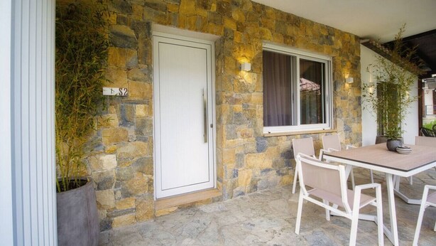 A stone exterior wall with a white door, a window with curtains, and a table with chairs. - Coel Luxury Suites S2 by RentalsPro (Nea Moudania)
