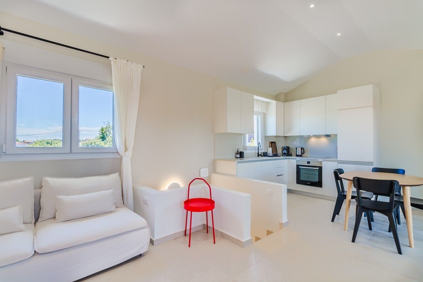 A modern, minimalist living room with a white sofa, a red chair, and a dining area with a table and chairs.