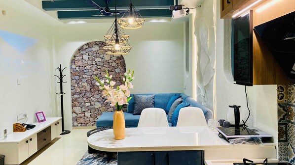 A modern living room with a stone wall, a blue sofa, a white dining table, and a ceiling fan. - Global Village BnB (Indore)