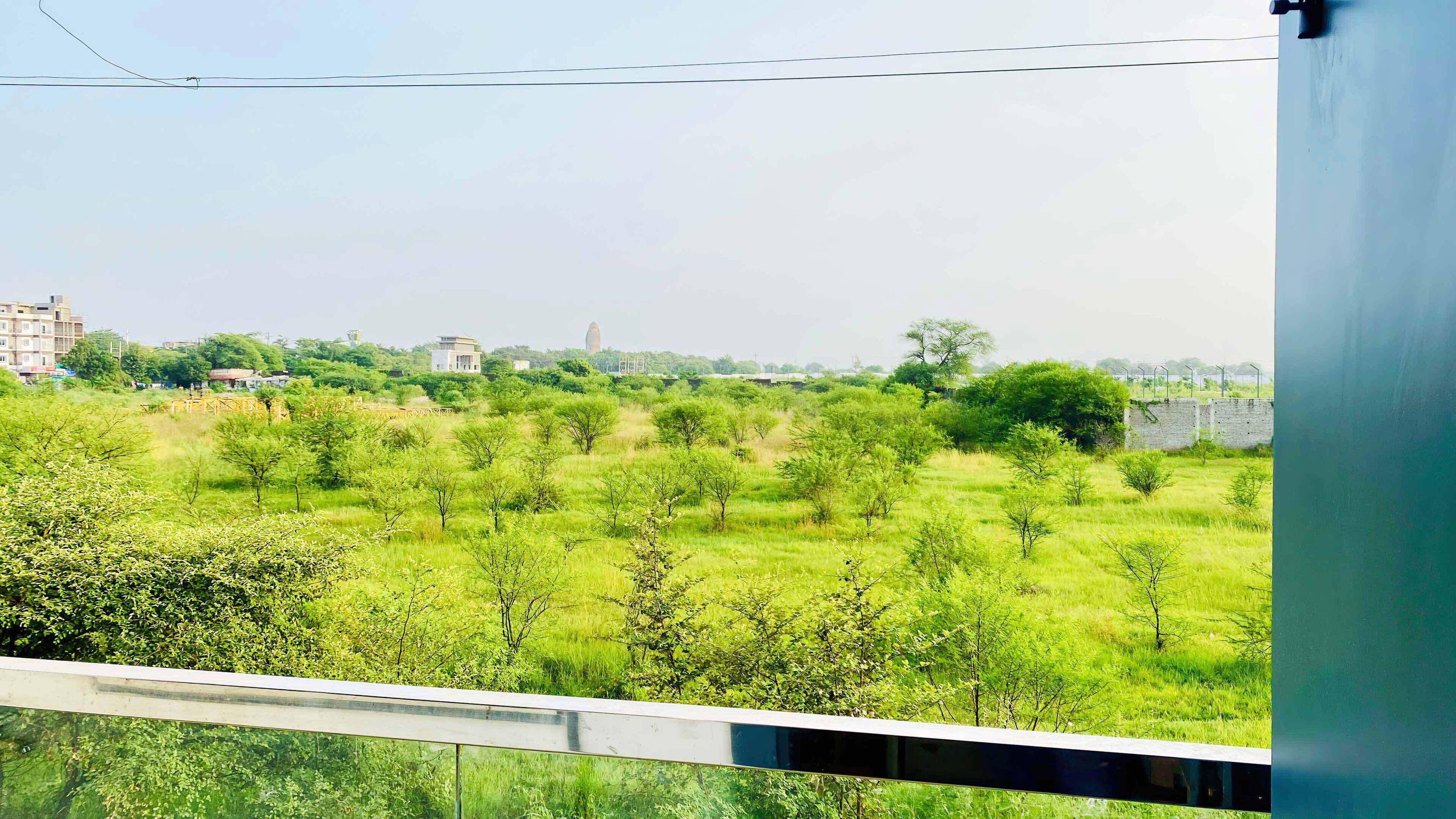 A view from a balcony overlooking a grassy field with trees, a distant building, and a clear sky.
