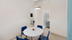 Un comedor moderno con una mesa redonda y sillas azules, una lámpara colgante y vista hacia un baño con ducha de vidrio.