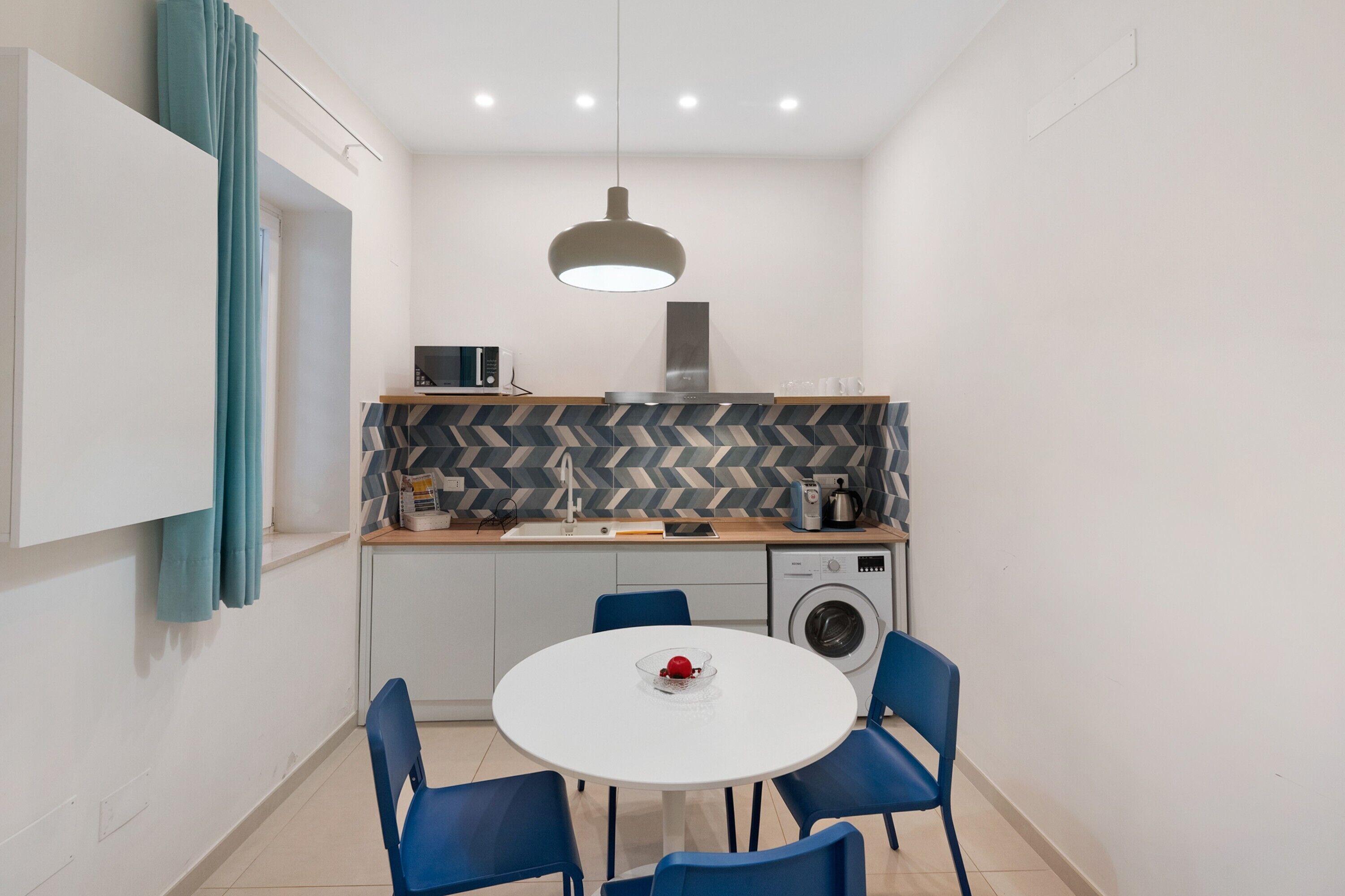 Une cuisine compacte équipée d’un four à micro-ondes, d’un grille-pain et d’un lave-linge. On y trouve une table à manger avec des chaises et un luminaire suspendu.