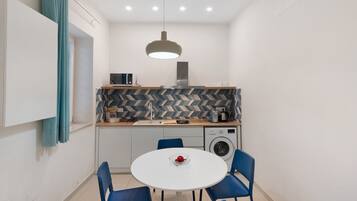 A compact kitchen with a microwave, toaster, and washing machine. There's a dining table with chairs and a hanging light fixture.
