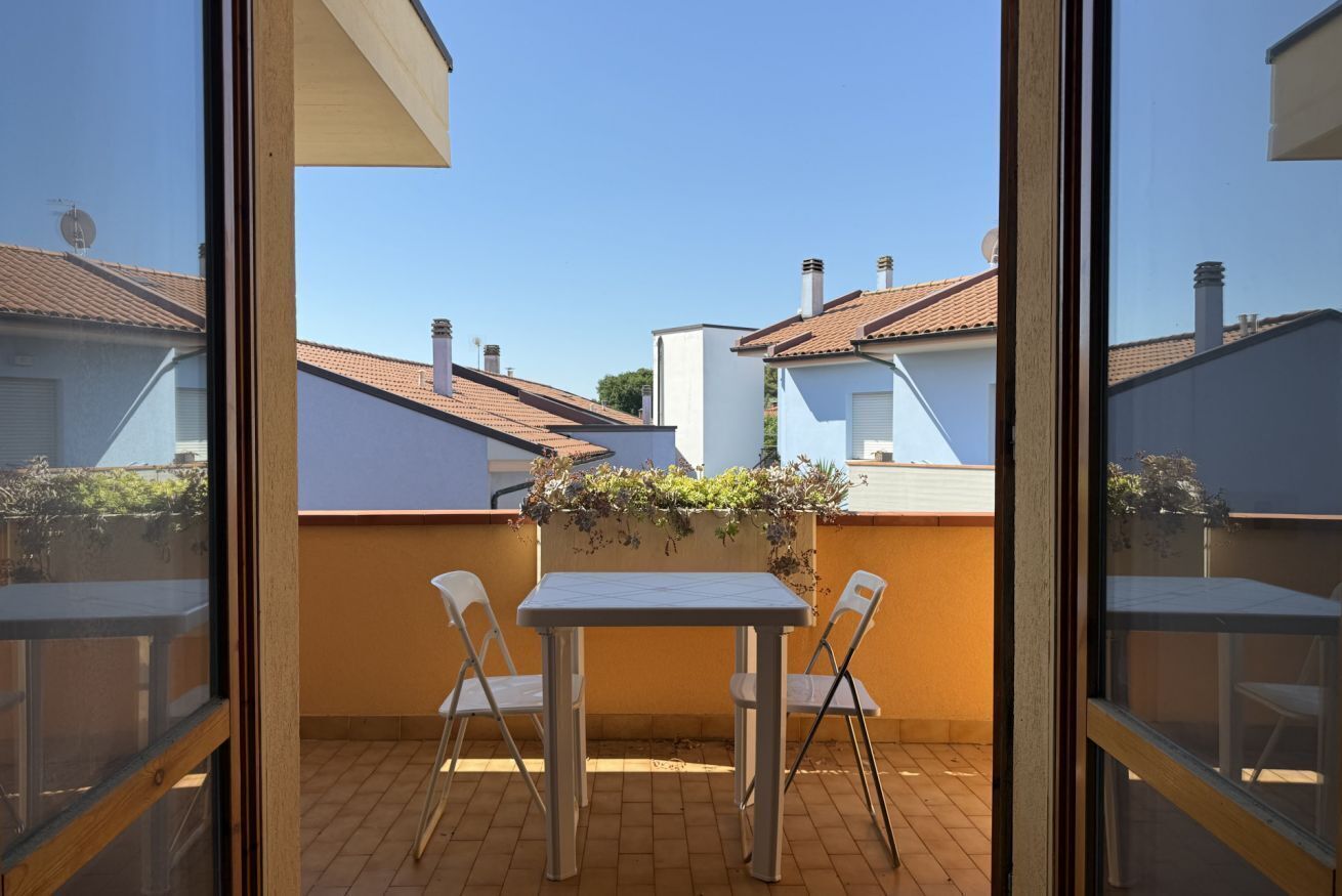 En balkon med et bord og stole, udsigt til nabobygninger og en klar himmel.