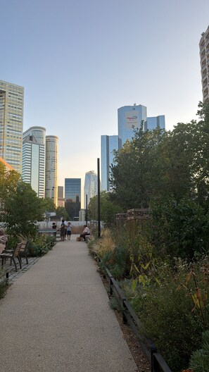 Un sentier qui traverse un parc agrémenté de bancs et de verdure, et est bordé d’immeubles modernes.