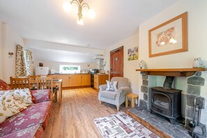 A cozy living room with a fireplace, wooden furniture, and a kitchen in the background. - Waters Edges cottage up to 12 in the beautiful peaceful Lake District location. (Selside)