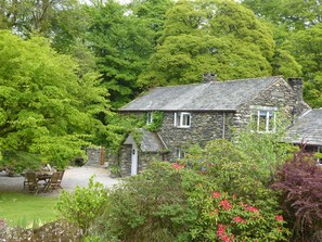 A stone house with a thatched roof, surrounded by lush greenery and flowering plants. - Waters Edges cottage up to 12 in the beautiful peaceful Lake District location. (Selside)