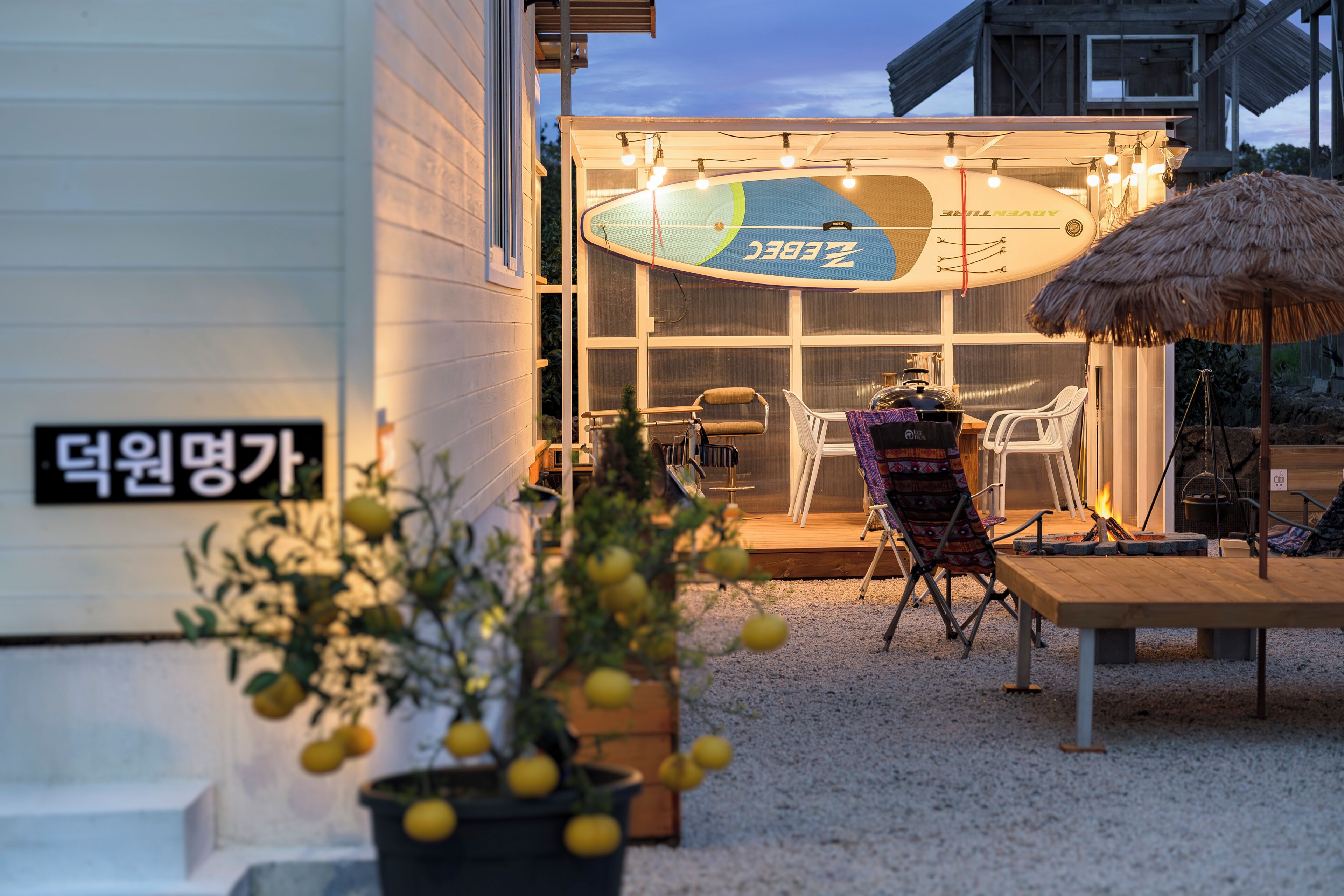 Un edificio con un letrero en coreano, una decoración de tabla de surf, mesas al aire libre y un parasol de paja.